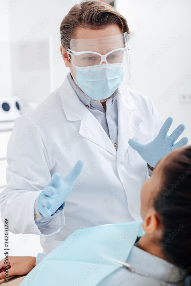 selective focus of dentist in face shield, medical mask and latex gloves gesturing near african american woman selective focus of dentist in face shield, medical mask and latex gloves gesturing near african american woman