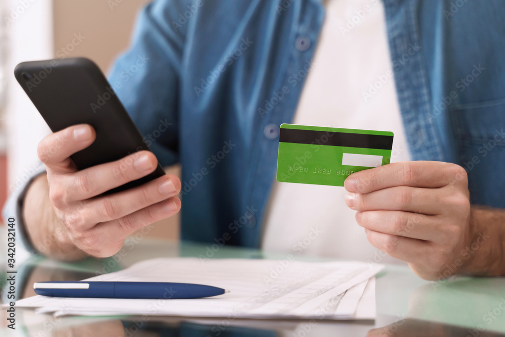 Internet banking, e-commerce and online trading concept. Closeup of young caucasian man holding mobile phone and credit card while paying bills at home Internet banking, e-commerce and online trading concept. Closeup of young caucasian man holding mobile phone and credit card while paying bills at home