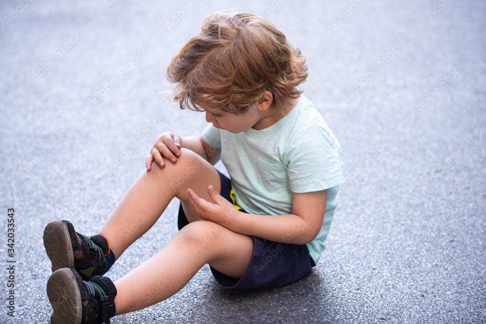 Little boy fell and scratched his knee, it hurts the wound and it keeps a handle on the leg, but not crying. Active boy, loves moving. A child sits on the road with a knee injury. Little boy fell and scratched his knee, it hurts the wound and it keeps a handle on the leg, but not crying. Active boy, loves moving. A child sits on the road with a knee injury.