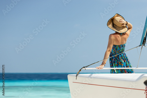 Back view of joyful attractive amazing woman in blue dress and hat enjoying azure water on the deck of a sailing yacht. Concept of sailing regatta, luxury vacation at sea.