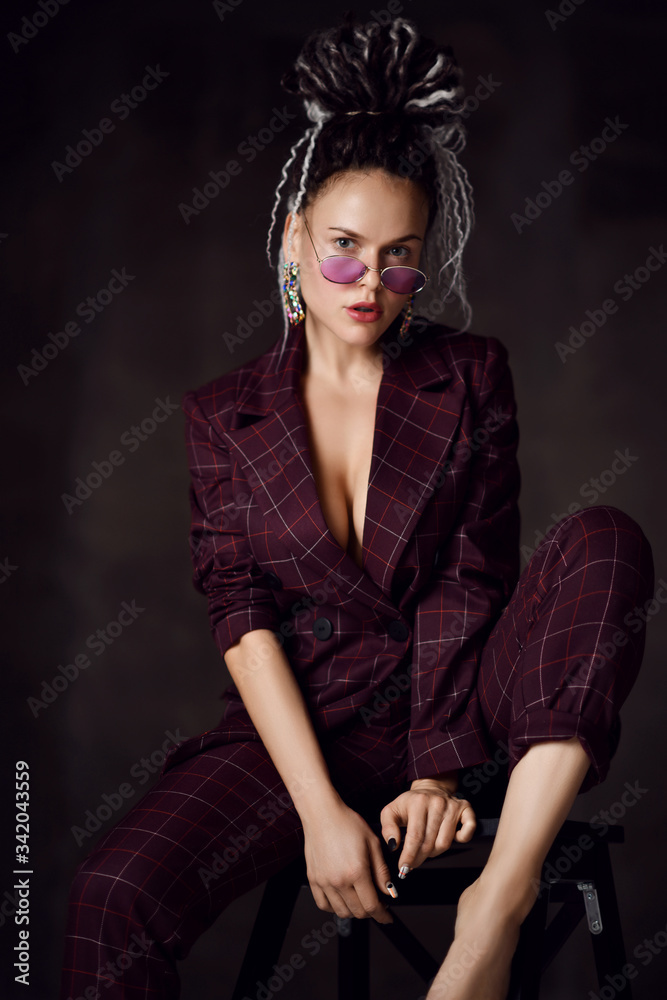 Young beautiful woman with dreadlocks hairstyle in sexy checkered suit with deep neckline sitting barefoot on chair Young beautiful woman with dreadlocks hairstyle in sexy checkered suit with deep neckline sitting barefoot on chair