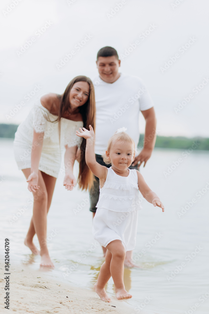 Mom, dad hugging daughter walking on the beach near lake. The concept of summer holiday. Mother's, father's, baby's day. Family spending time together on nature. Family look. Sun light. Mom, dad hugging daughter walking on the beach near lake. The concept of summer holiday. Mother's, father's, baby's day. Family spending time together on nature. Family look. Sun light.