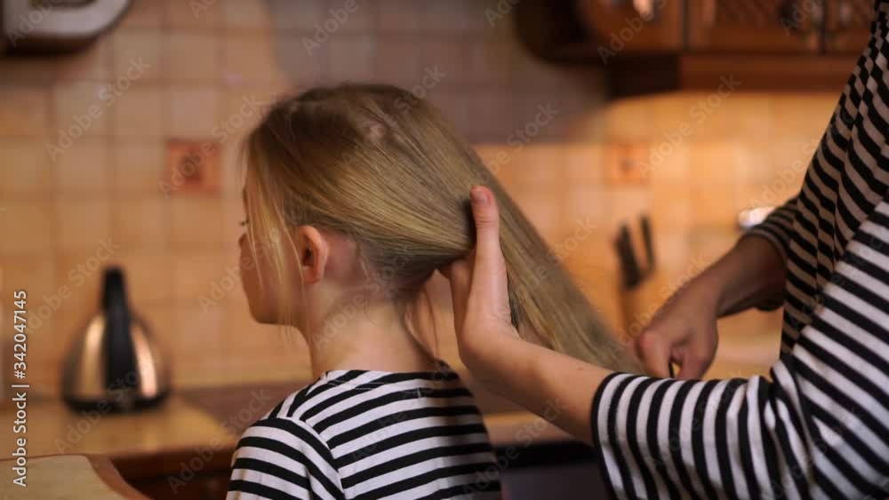 Mother brush comb daughter child long blond hair with colorful comb brush wearing striped dress at cosy wooden kitchen home Mother brush comb daughter child long blond hair with colorful comb brush wearing striped dress at cosy wooden kitchen home