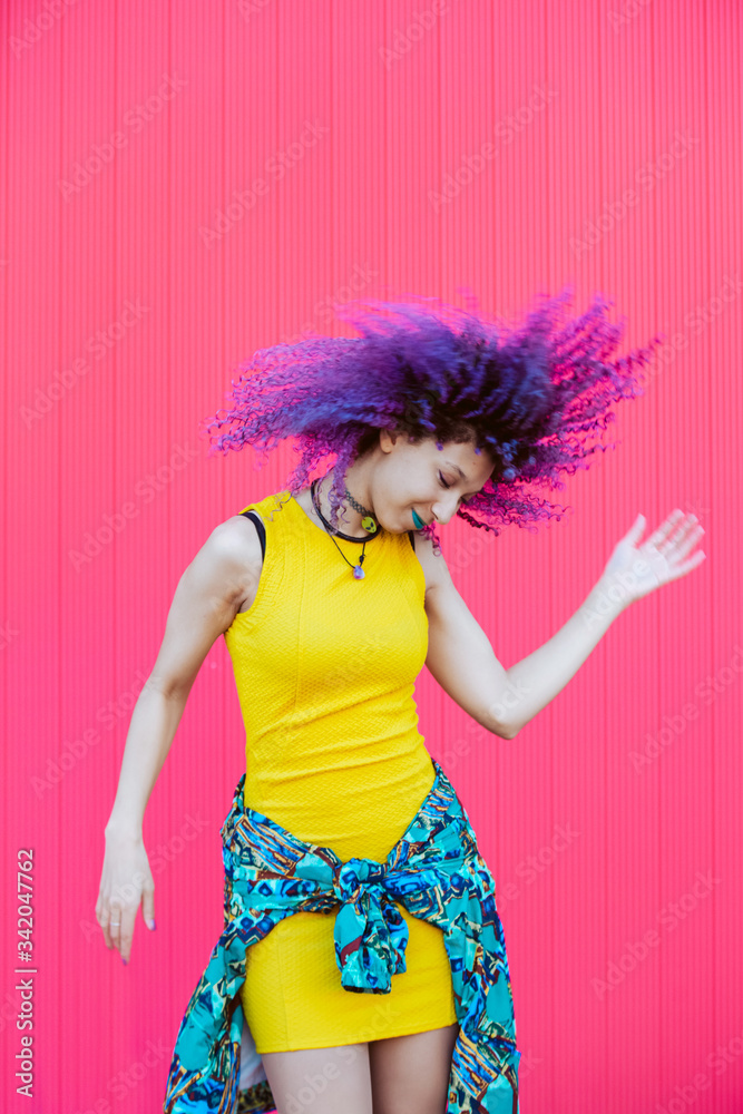 Millennial woman with bright yellow dress isolated on pink background. Hair dyed with psychedelic bright colors concept. Happy music leisure concept. Cheerful mixed raced woman dancing and having fun. Millennial woman with bright yellow dress isolated on pink background. Hair dyed with psychedelic bright colors concept. Happy music leisure concept. Cheerful mixed raced woman dancing and having fun.