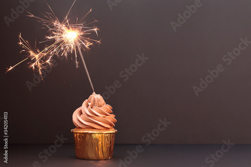A chocolate cupcake in a golden cup with a Sparkler candle. Black background
