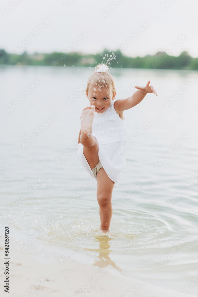 Naklejka premium cute baby girl walking and having fun on the beach near lake. The concept of summer holiday. baby's day. Family spending time together on nature.