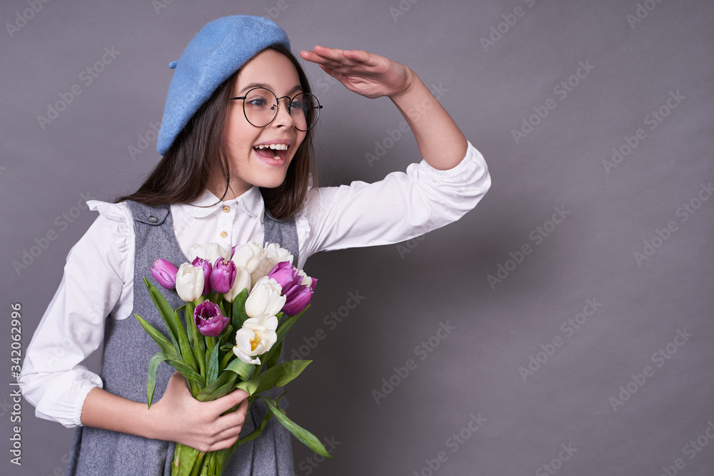 Cute fashionable emotional lady in a stylish blue hat in a beret and glasses holds flowers tulips in a gray background. Cute fashionable emotional lady in a stylish blue hat in a beret and glasses holds flowers tulips in a gray background.