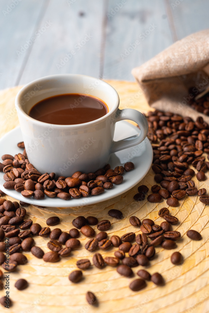 White cup and saucer with coffee and surrounded by roasted coffee beans on a wooden table and a bag White cup and saucer with coffee and surrounded by roasted coffee beans on a wooden table and a bag