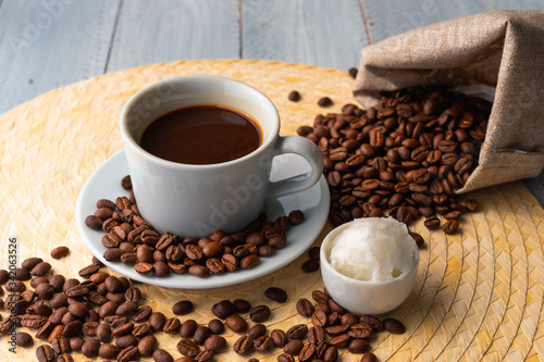 White cup and saucer with coffee and surrounded by roasted coffee beans on a wooden table and a bag next to a small cup of coconut oil