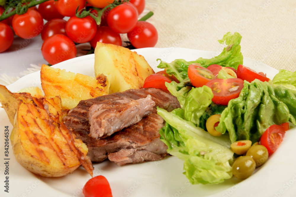pieces of grilled steak next to potatoes grilled next to lettuce with tomatoes and olives on a white plate