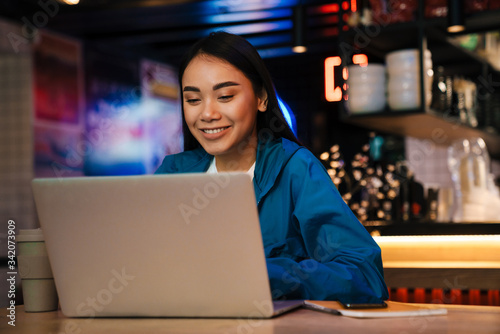 Photo of cheerful asian woman working with laptop and smiling in cafe