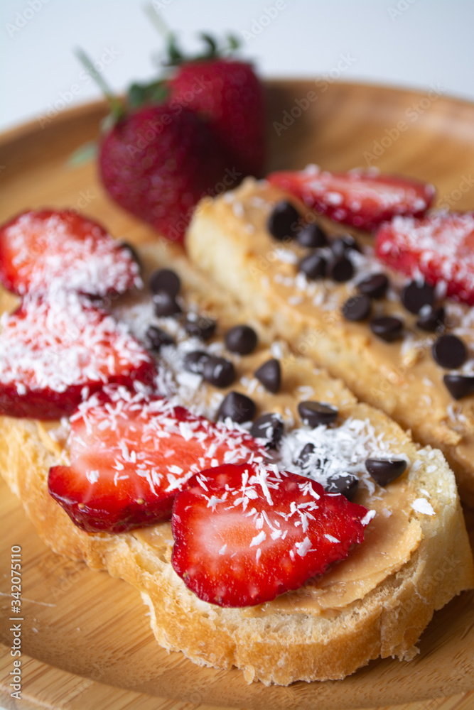 Peanut butter spread toasts with chocolate chips, strawberries, coconut flakes and homemade bread on a wooden plate