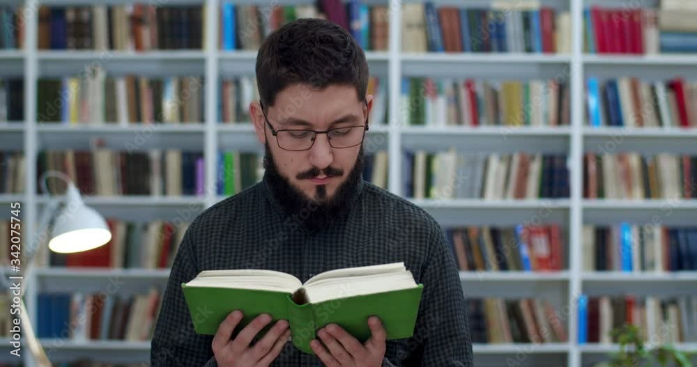 Young handsome Caucasian man in glasses and with beard reading textbook in library. Close up of male student or scientific researcher with book in hands studying in public bibliotheca. Study concept. Young handsome Caucasian man in glasses and with beard reading textbook in library. Close up of male student or scientific researcher with book in hands studying in public bibliotheca. Study concept.