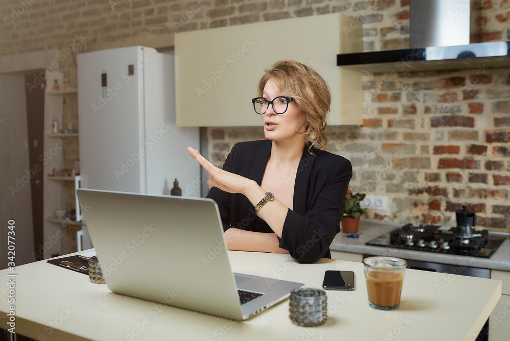 A woman in her kitchen works remotely on a laptop. A blond girl in glasses gesturing while talking to her colleagues in a video call at home. A woman in her kitchen works remotely on a laptop. A blond girl in glasses gesturing while talking to her colleagues in a video call at home.