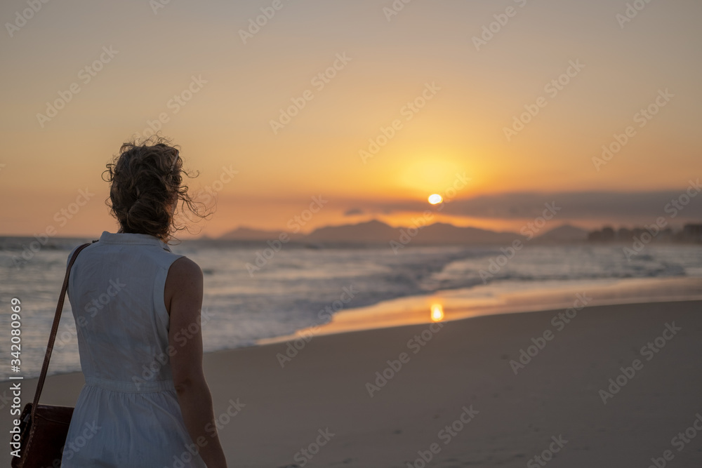 Fototapeta premium Girl in a light dress at sunset on the beach against the background of the ocean mountains and sun