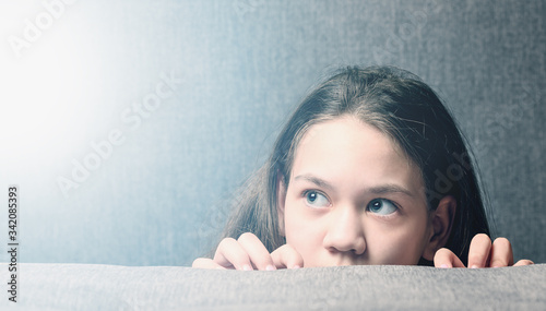 Frightened girl with big eyes peers out from behind the sofa. Concept child abuse.