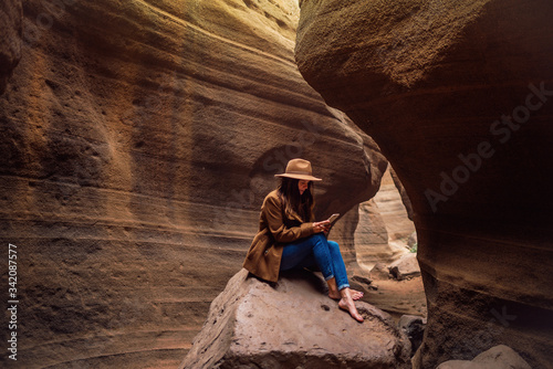 Side view of stylish young barefooted female traveler in hat using smartphone while sitting on stone in rocky ravine and resting after hiking photo