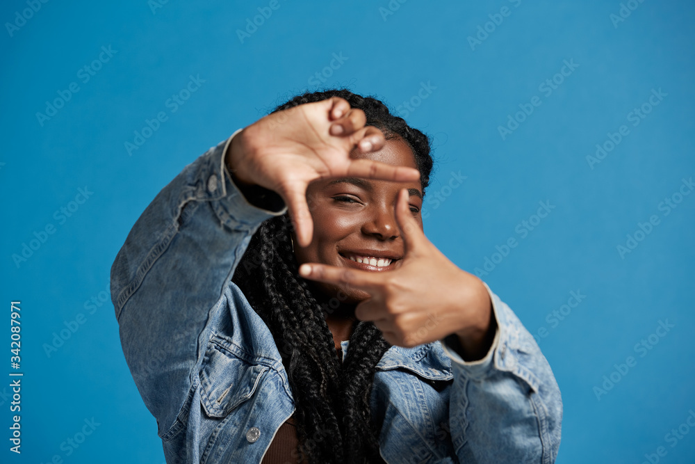 Happy black female with braids in denim jacket smiling and looking at camera while framing face with hands against blue background Happy black female with braids in denim jacket smiling and looking at camera while framing face with hands against blue background