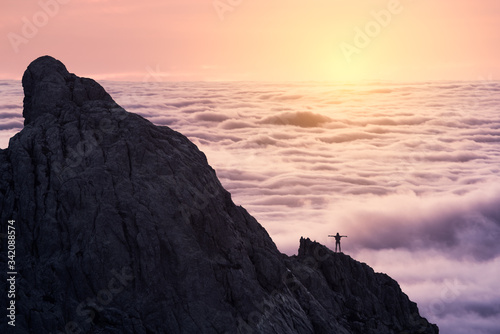 Silhouette of unrecognizable person stretching arms standing on peak of rough cliff with colorful clouds on background during sunset photo