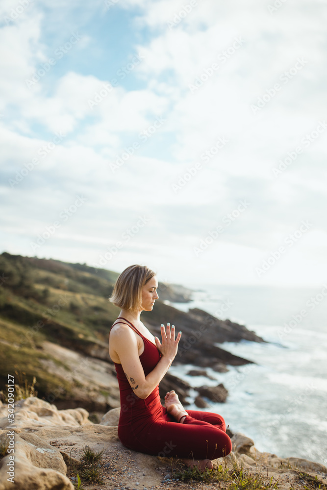 Side view of woman with closed eyes in red sporty wear working out and practicing lotus pose yoga with prayer hands on empty rocky beach Side view of woman with closed eyes in red sporty wear working out and practicing lotus pose yoga with prayer hands on empty rocky beach