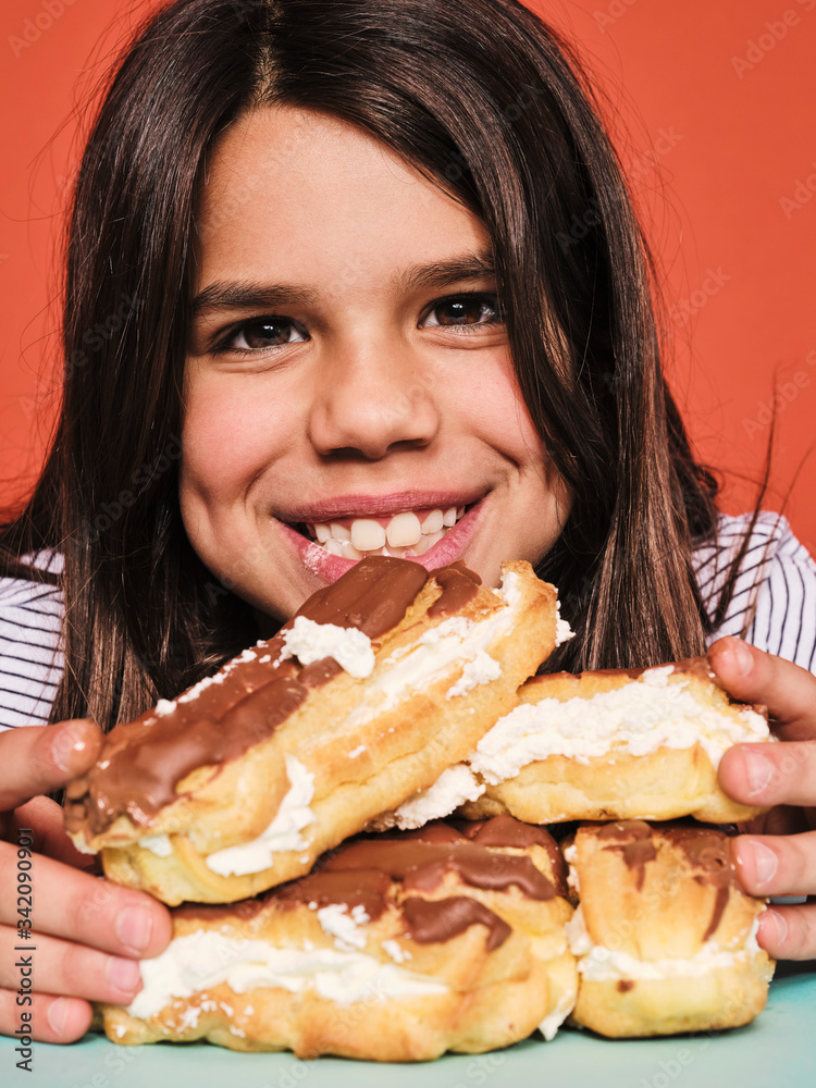 Closeup portrait of cheerful little girl in casual wear enjoying sweet eclairs with chocolate while sitting at table against red background Closeup portrait of cheerful little girl in casual wear enjoying sweet eclairs with chocolate while sitting at table against red background