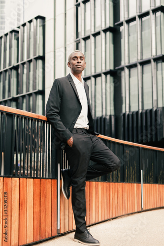 Full body positive African American manager with hand in pocket of suit leaning on fence and looking away on city street