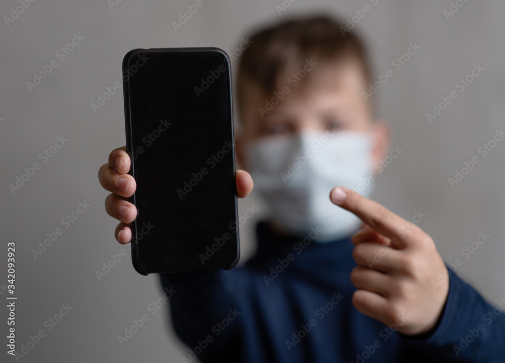 A boy in a medical mask holds a cell phone in his hands and points his finger at the phone. Coronovirus Pandemic Concept