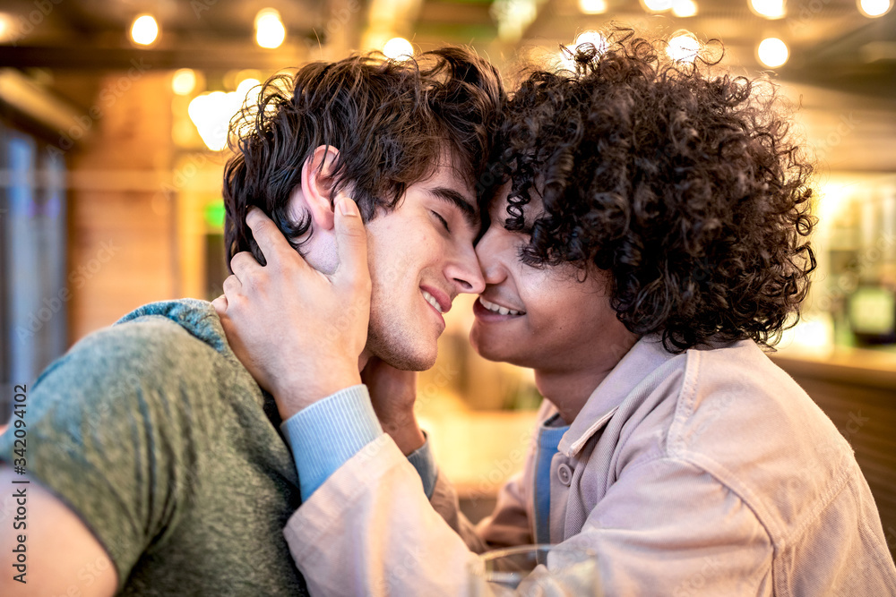 Side view of excited ethnic men embracing each other with closed eyes on table and laughing during romantic date in modern cafeteria Side view of excited ethnic men embracing each other with closed eyes on table and laughing during romantic date in modern cafeteria