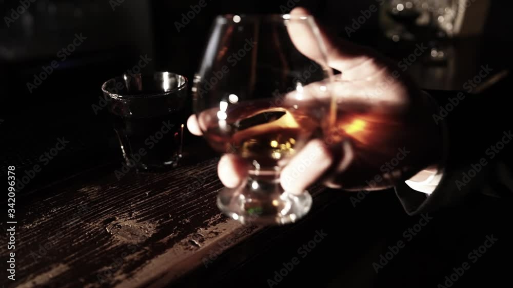 A glass of whiskey stands on a wooden bar counter that a man takes in a dark bar. A glass of whiskey stands on a wooden bar counter that a man takes in a dark bar.