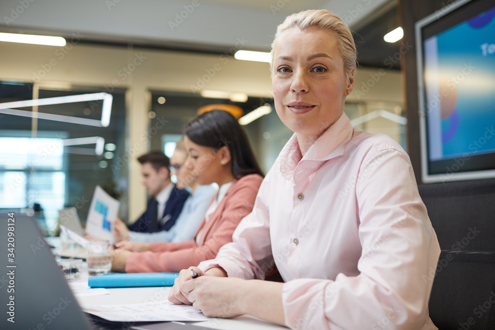Naklejka premium Portrait of mature businesswoman with short hair looking at camera while working at the table on laptop with her colleagues in the background