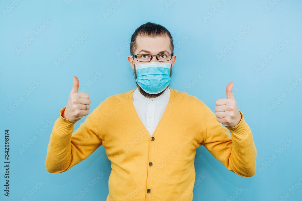 A young man in a medical mask on a blue background. A man in a yellow cardigan shows a hand gesture OK. We're going to beat coronavirus. The COVID-2019 epidemic. Positive boy. A young man in a medical mask on a blue background. A man in a yellow cardigan shows a hand gesture OK. We're going to beat coronavirus. The COVID-2019 epidemic. Positive boy.