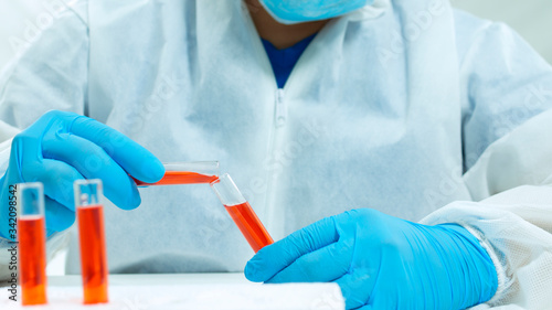 Latin doctor in bioprotective suit, face mask and blue gloves mixing a red liquid between two glass test tubes on white background photo