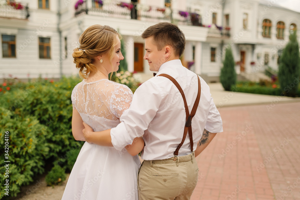 Stylish groom and happy bride are hugging and looking at each other on the background of architecture. Wedding day.