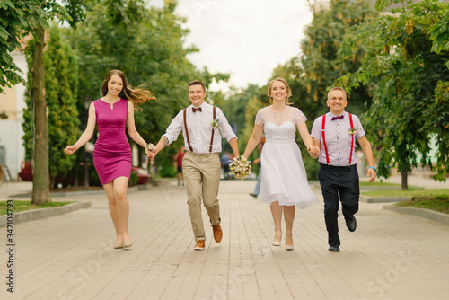 Newlyweds are have fun at wedding day together with their friends, holding hands they run along the street.