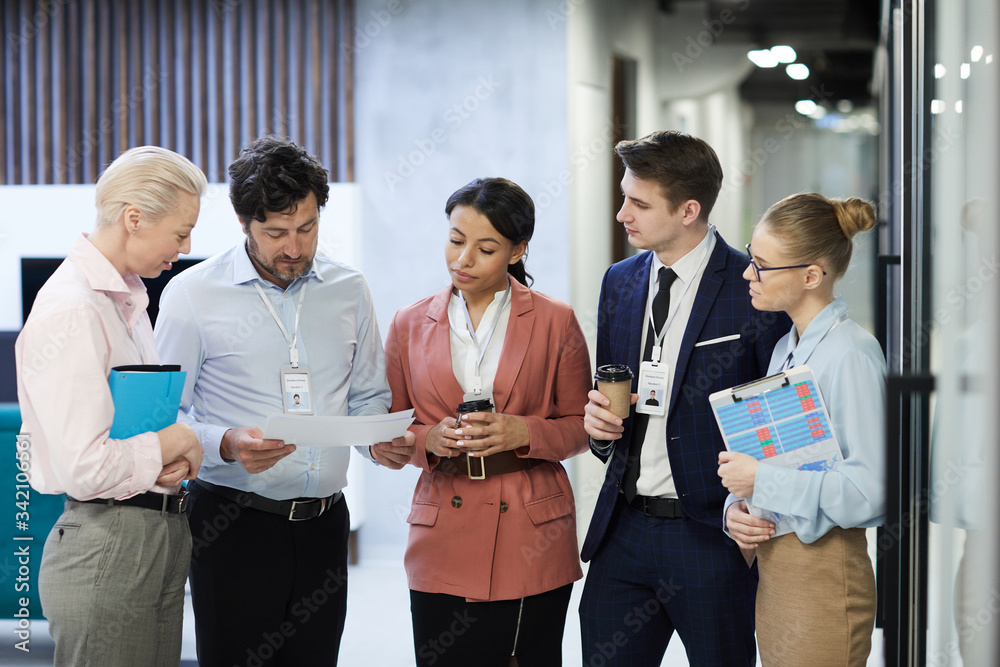 Group of business people standing and discussing documents they preparing for the meeting while standing at office corridor Group of business people standing and discussing documents they preparing for the meeting while standing at office corridor