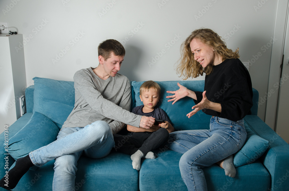 Child with a phone, parents swear in quarantine. Young family, a man, a woman and sons are sitting in a room on a blue sofa. Child with a phone, parents swear in quarantine. Young family, a man, a woman and sons are sitting in a room on a blue sofa.