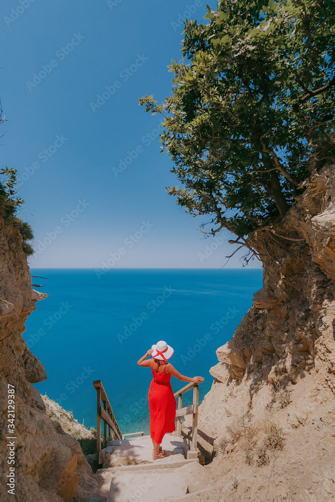 girl in a red dress and a white hat stands on the steps against the sea