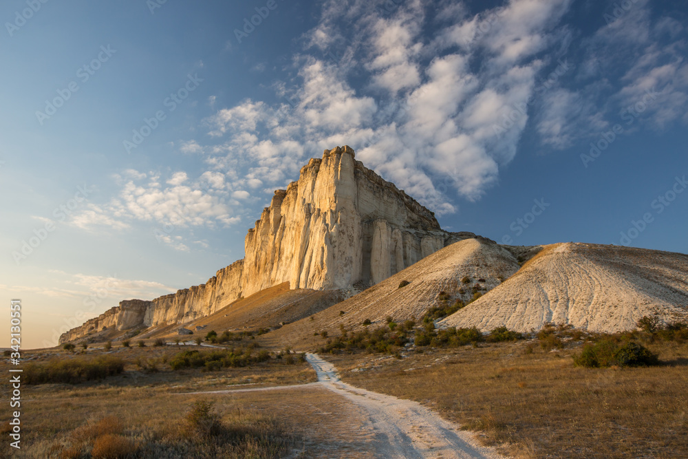 Obraz premium White rock against a beautiful sunset, light clouds fly over the rock and the road leads to the foot of the rock.