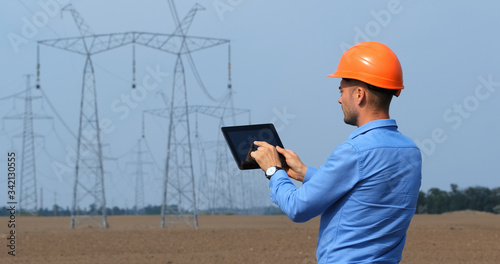 An engineer in an orange safety helmet works in a tablet against the background of Electrical supports and power lines.