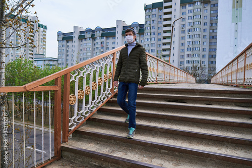 teen boy walks down the street during the day, a pedestrian walkway and high-rise buildings with apartments, a residential area, a medical mask on his face protects against viruses and dust © soleg