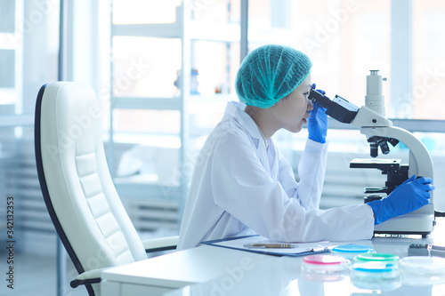 Female doctor in protective gloves and cap examining analysis on microscope while sitting at her workplace at office