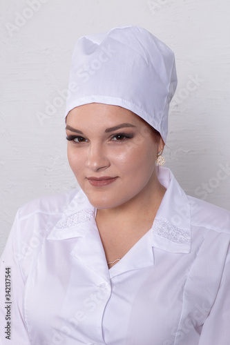 Portrait of attractive young female doctor in white medical jacket and white medical cap on a grey background.