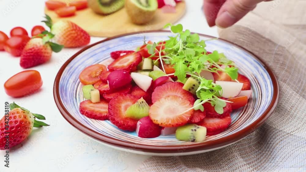 Vegetarian fruits and vegetables salad of strawberry, kiwi, tomatoes, on white concrete background and linen textile decorated with microgreen sprouts by male hand. Side view, close up.