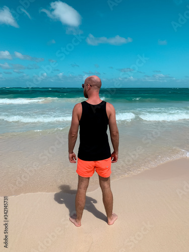 European hairless bearded man male guy standing on the beach in black sleeveless shirt singlet with empty space for text or print on ocean sea background. View from back