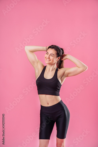 Slim pumped up girl fitness trainer stands against pink background in the studio in black sportswear. She hugs herself with hands, closing her eyes, smiling, dreaming, and raising her head up