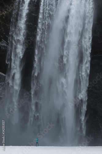 Person in blue jacket standing in front of massive waterfall Sk  gafoss  Iceland