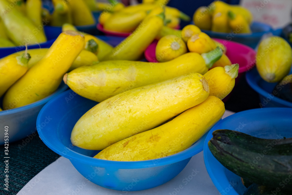 Yellow squash in blue bowls and green zucchini on a table at a farmer's market. The squash has a flat bottom and tappers along its long body at the neck. The small vegetables are laid in small bowls.