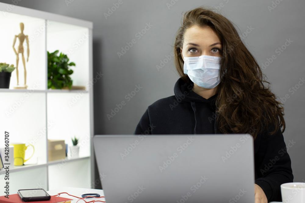 Young beautiful business woman with long hair in medical protective mask looking seriously at camera working at computer from home office during self-isolation. Covid-19 coronavirus quarantine concept Young beautiful business woman with long hair in medical protective mask looking seriously at camera working at computer from home office during self-isolation. Covid-19 coronavirus quarantine concept