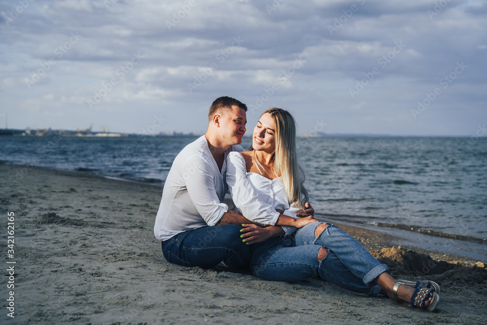 Lifestyle romantic evening for two. Young lovers went on a trip to the sea. Sitting on the sand in the evening sun and hugging, talking, kissing and enjoying the absence of people around.