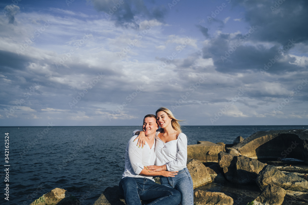 Fototapeta premium Two lovers blondes spending a romantic evening on the beach. They feeling evening sun, hugging, kissing and laughing, sitting on the worm stones. The girl is sitting on the arms of a guy.